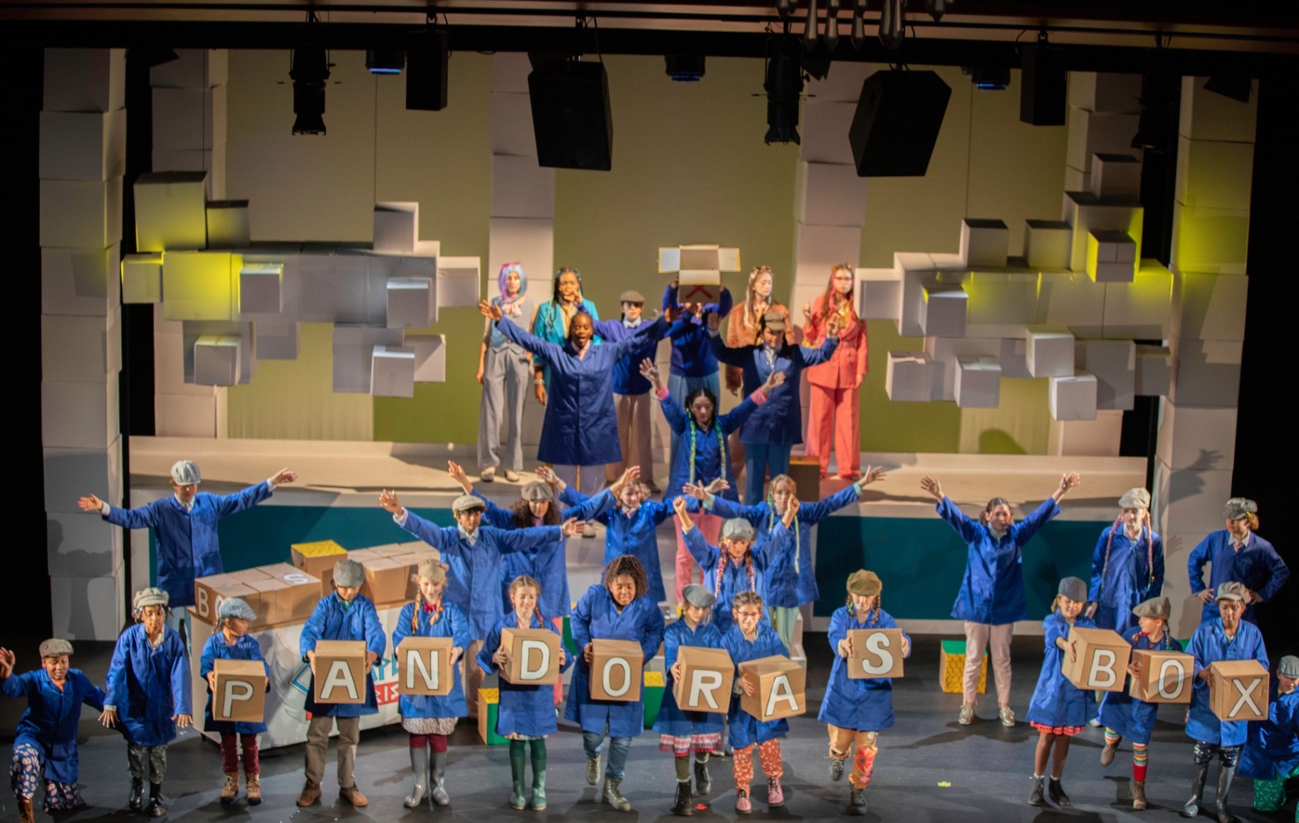 The full company assembled on a set of stacked white cardboard boxes, the mortals in blue overalls and flat caps holding letter blocks spelling 'PANDORA'S BOX' while the gods stand above them.