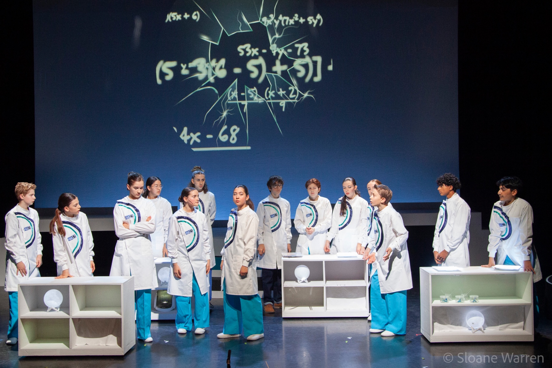 The ensemble of scientists in white coats stand deep in thought before a projected chalkboard of tangled equations, its surface cracked as though shattered.