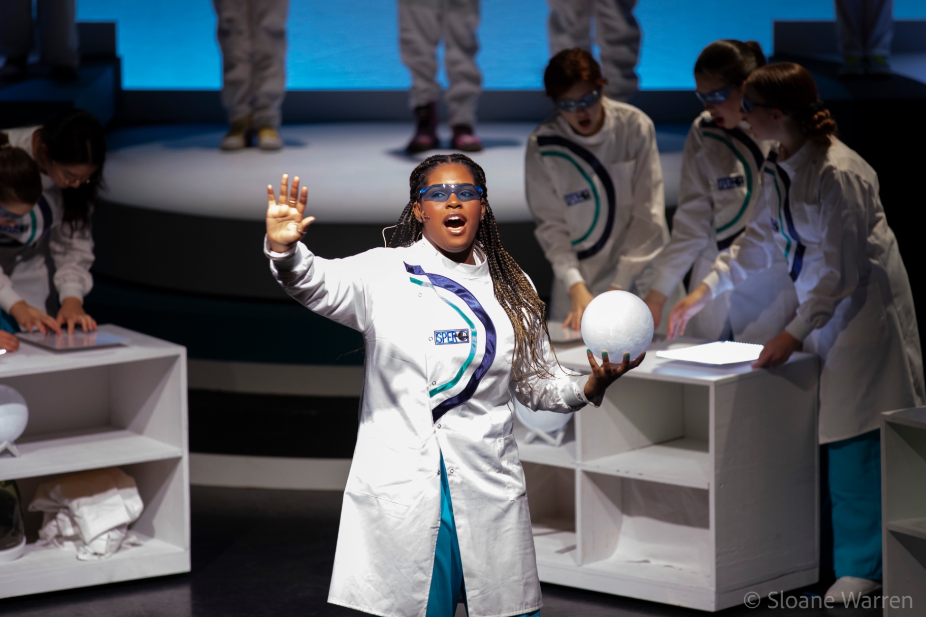 The lead scientist sings out to the audience, one hand raised, the other cradling a glowing white sphere as her colleagues work at their benches behind her.