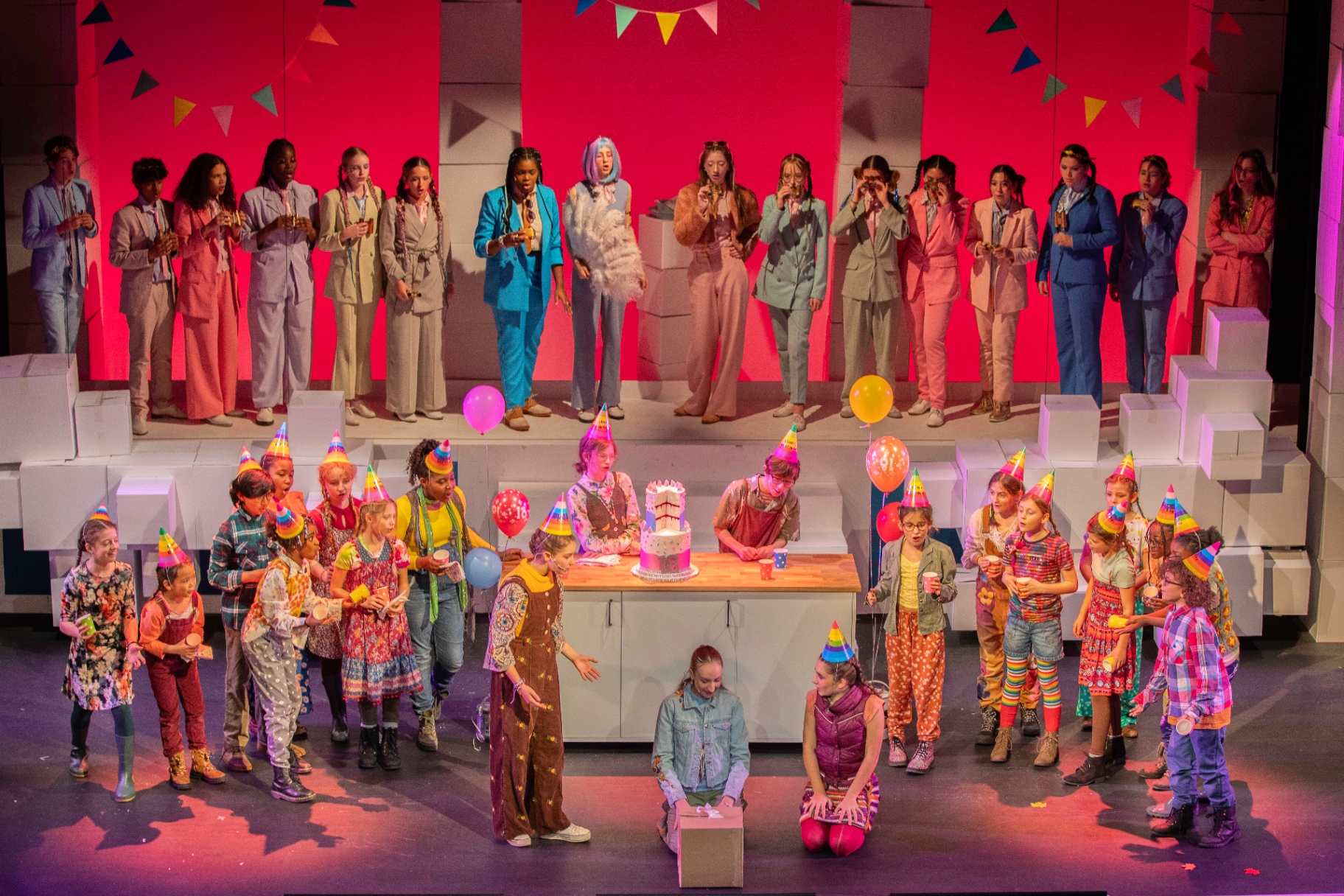 The full company of a London Youth Opera production on stage together - chorus in pastel suits raised above, younger performers in party hats gathered around a birthday cake below.