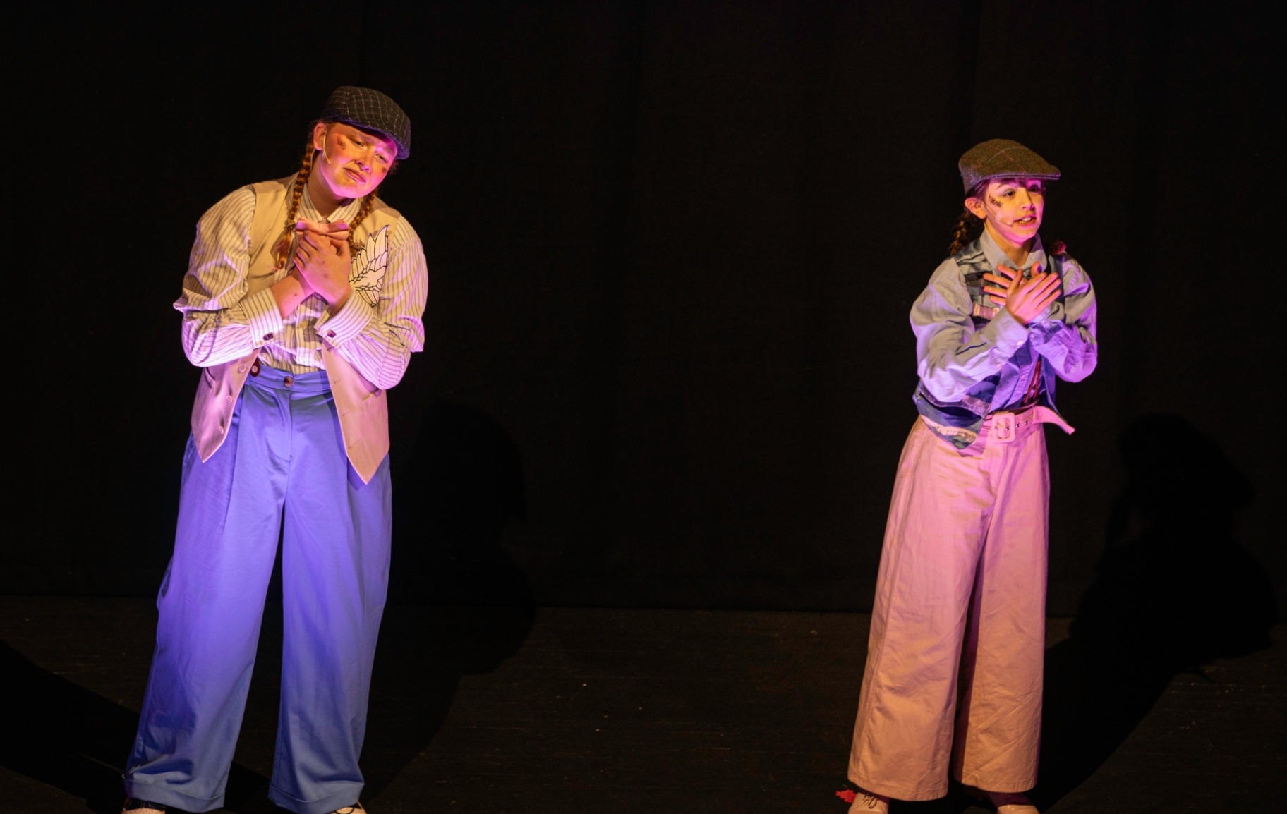 Two Olympus Enterprises messengers in flat caps and wide-legged trousers stand apart on a darkened stage, hands clasped to their chests under warm pink light.
