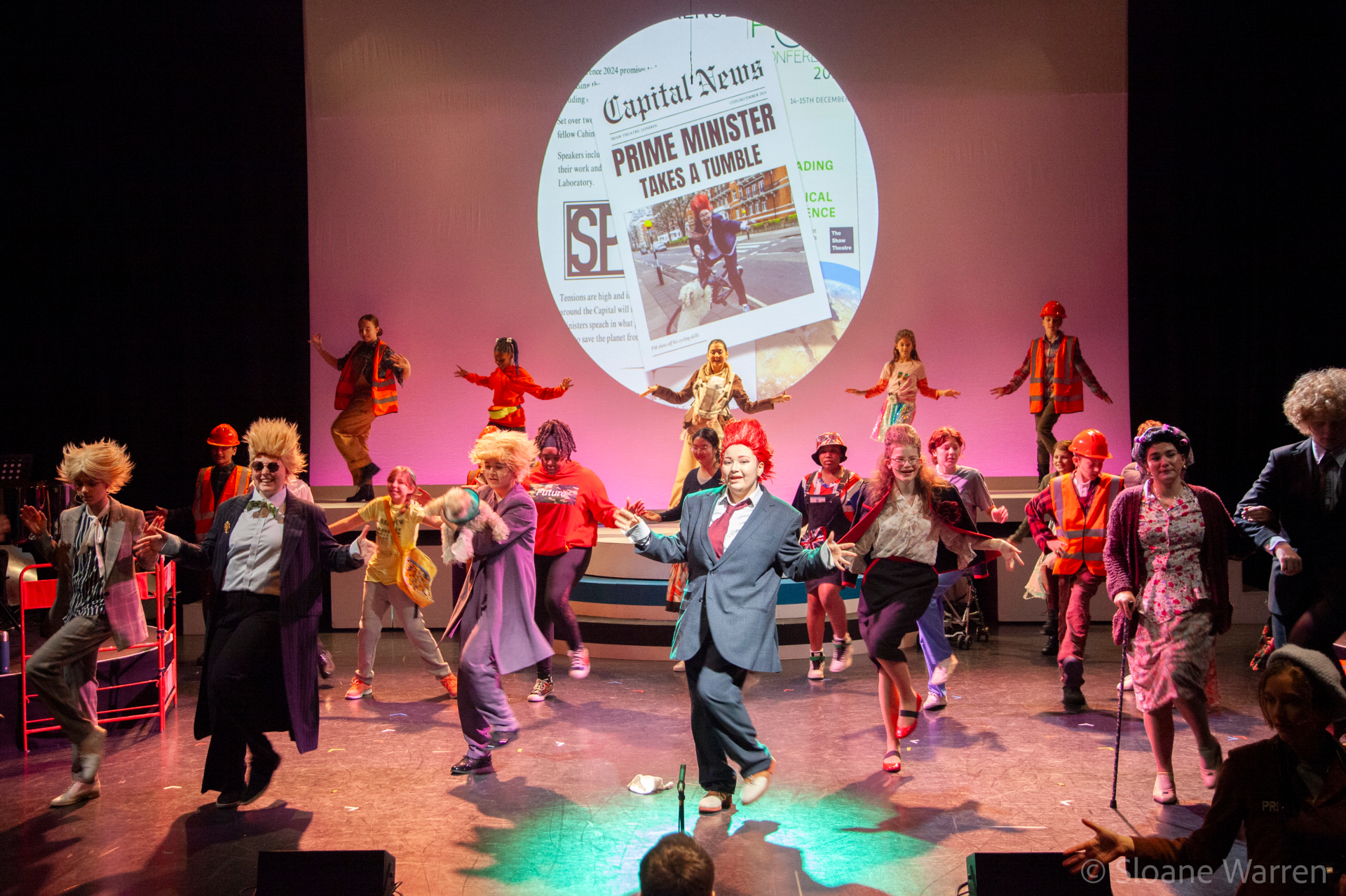 The Prime Minister leads the full company in a chaotic, joyful dance break, a projected newspaper headline reading 'Prime Minister Takes a Tumble' behind them.