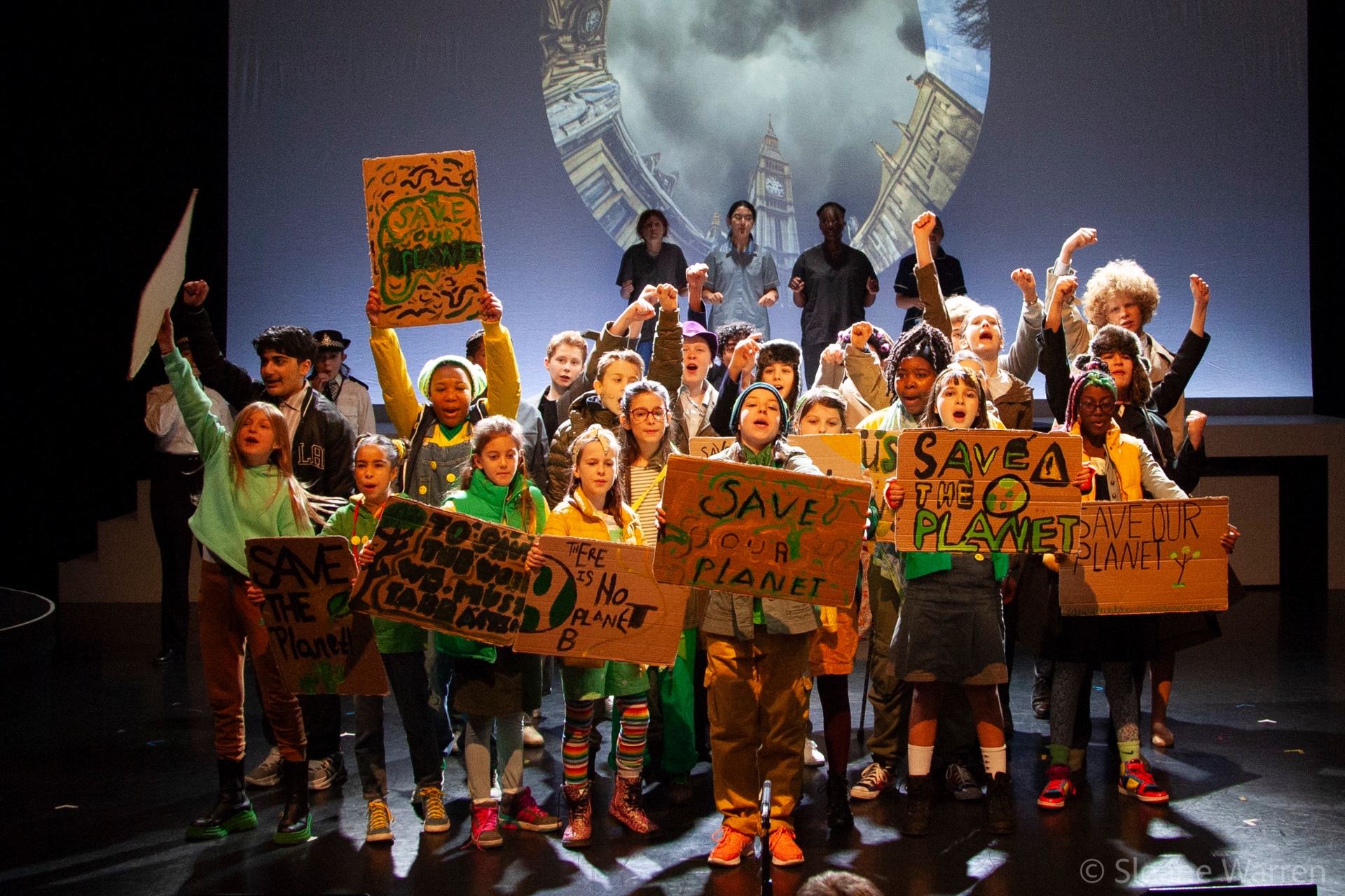The full company at the climax of the protest scene, fists raised and hand-painted placards held high, a projected image of Big Ben looming behind them.