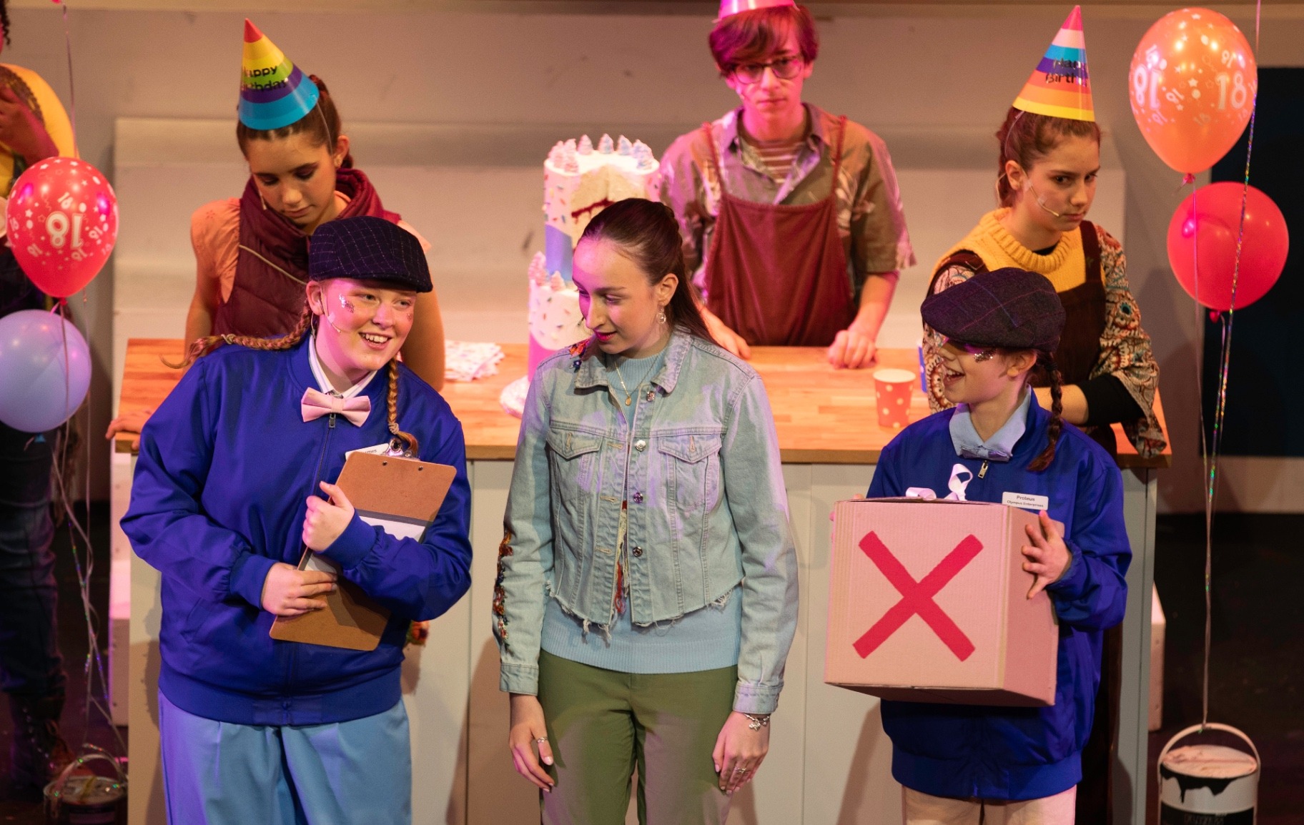 Pandora receives a plain cardboard box marked with a red X from two Olympus Enterprises messengers, her family gathered around a birthday cake behind her.