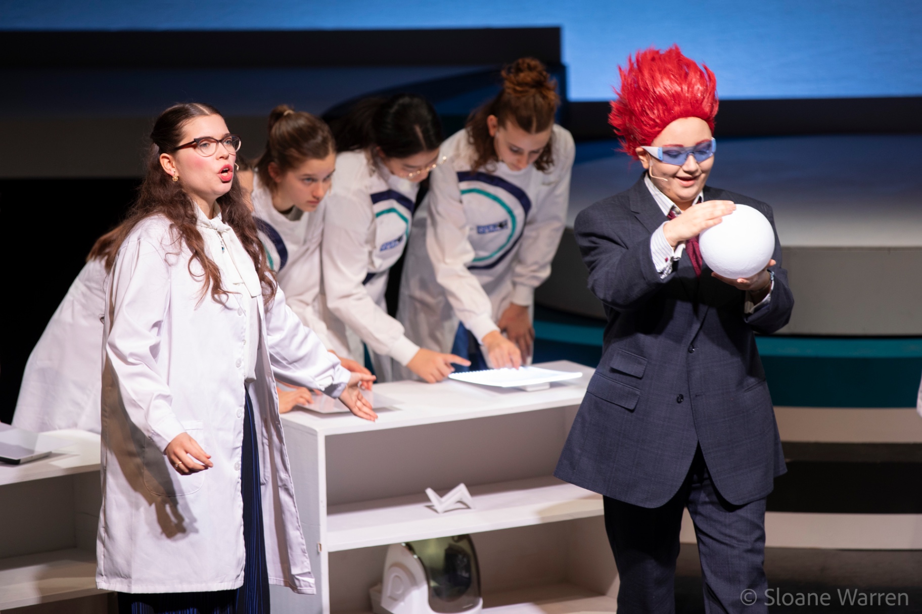 The red-haired Prime Minister holds a small white globe aloft while a scientist stands ready to respond, a chorus of colleagues bent over their workbench between them.