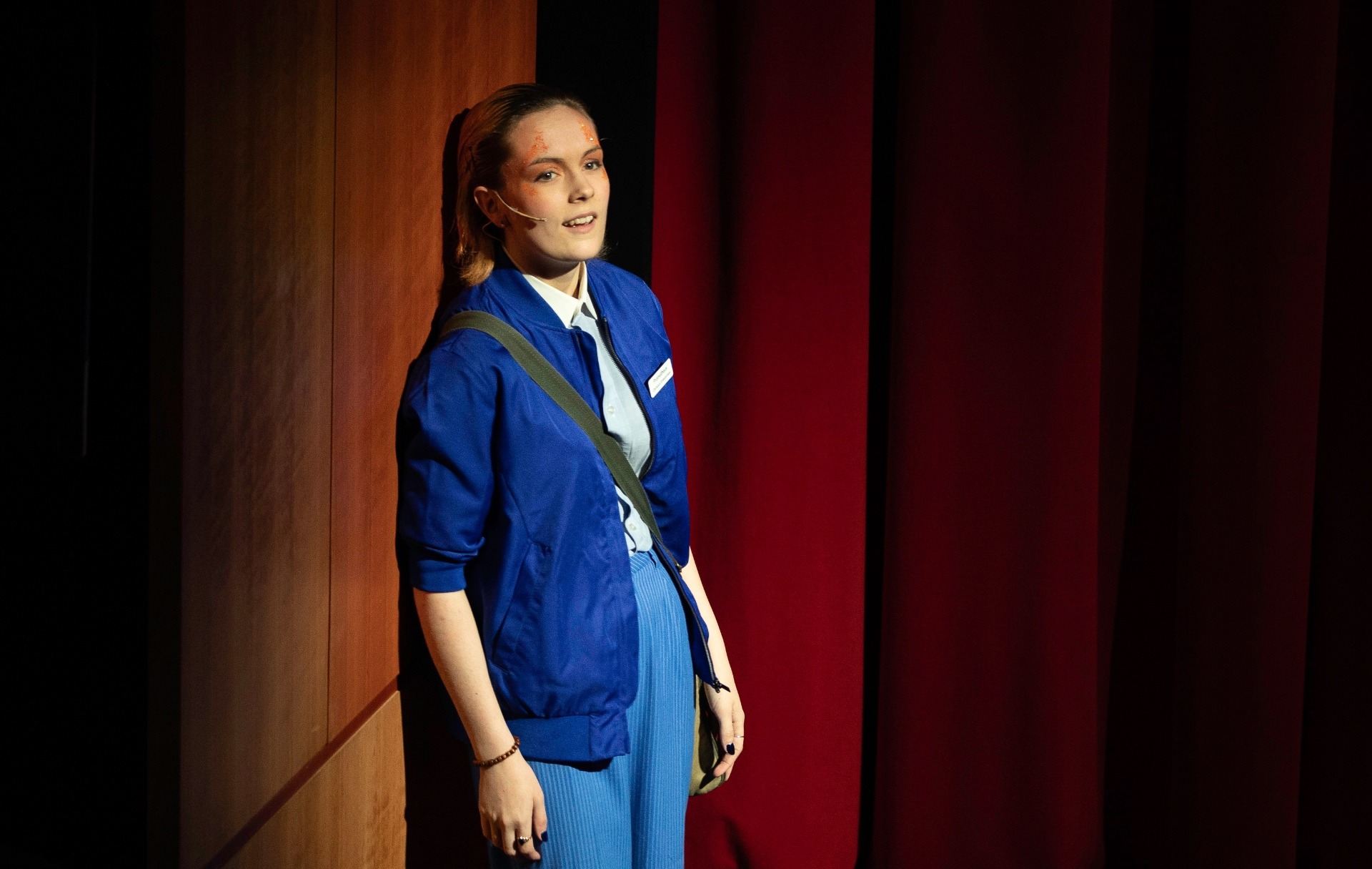 Prometheus, in an Olympus Enterprises blue bomber jacket and messenger bag, stands lit against a red curtain and wooden panelled backdrop.