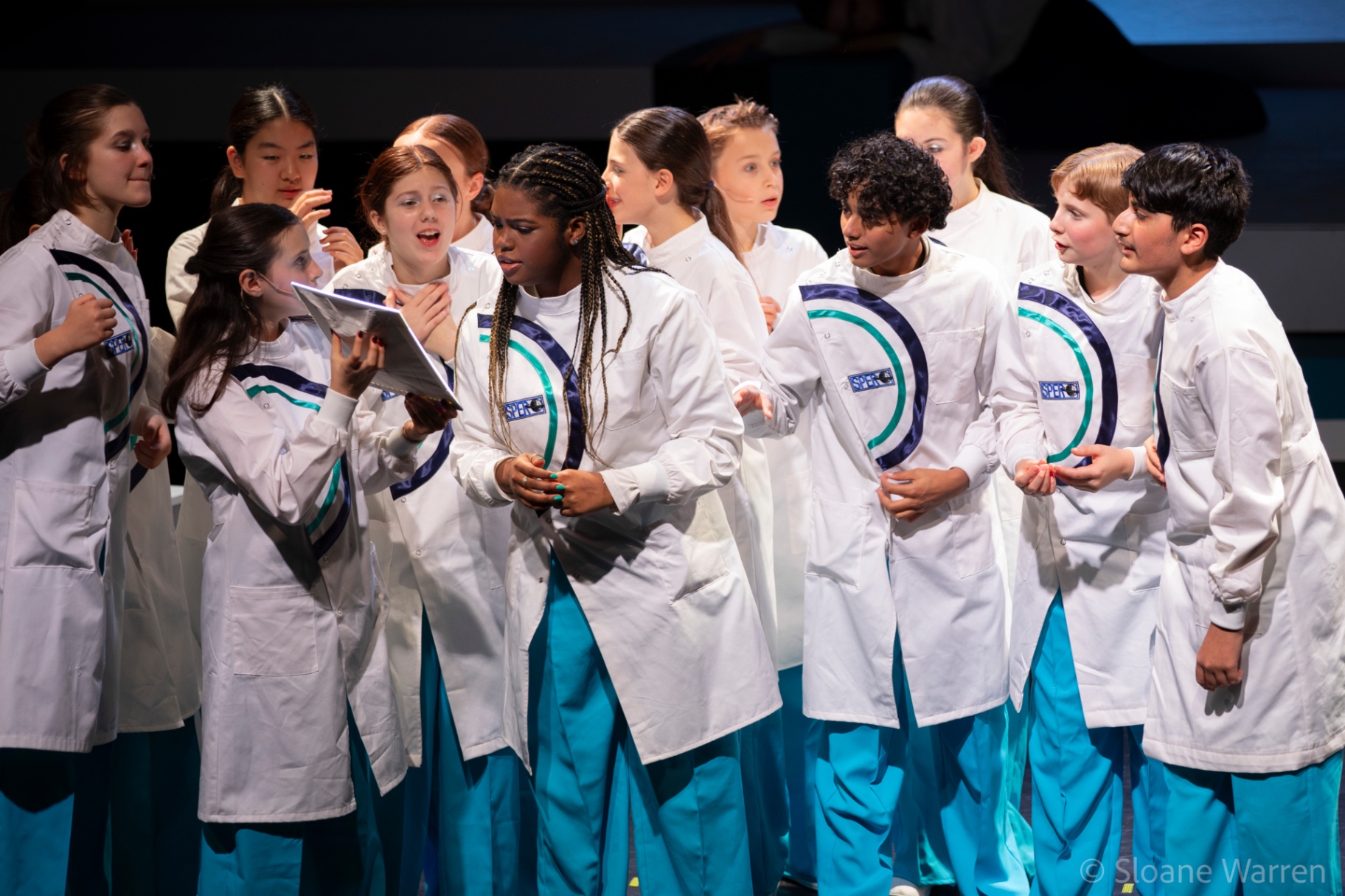 A cluster of young scientists lean in around a tablet, faces animated with excitement and alarm as they read a breakthrough.